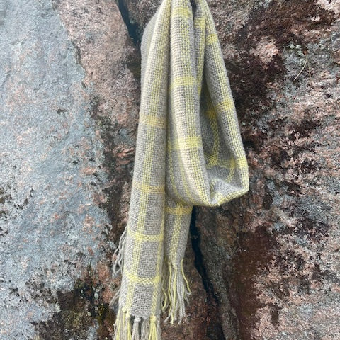 Kapea ruudullinen villahuivi – vaaleanharmaa & keltainen roikkumassakuten kaulassa. Kallio taustalla.