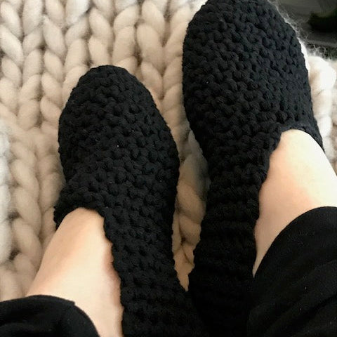 Mustat kotitossut henkilön jalassa, taustalla muhkea villaviltti.