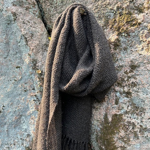 Tummanharmaa villahuivi hapsuilla roikkumassa kuten kaulassa kalliota vasten.